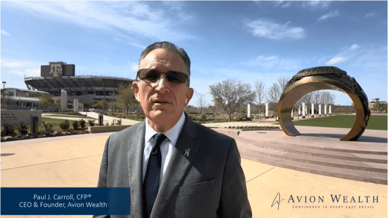 Man in suit near a large ring sculpture at a stadium, sunny day.