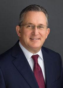 Smiling man in suit and tie, wearing glasses, against a neutral background.