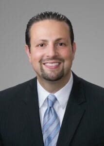 Smiling man in business suit with blue tie against gray background.