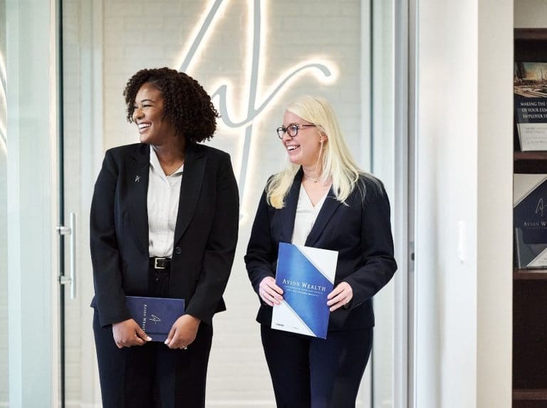 Two women in business attire hold Avion Wealth folders, smiling in an office. Welcoming guests for Expert Wealth Management Planning.